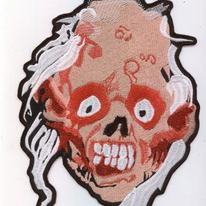 May include: Embroidered patch featuring a zombie face with red, white, and black details. The design includes exposed teeth, red eyes, and white hair-like elements. The patch has a black border and is likely for clothing or accessories.