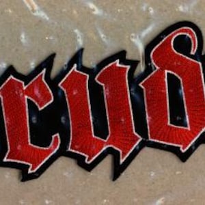 Könnte beinhalten: Ein gestickter Aufnäher mit dem Wort Drudkh in einer stilisierten Schriftart mit scharfen Kanten. Die Buchstaben sind leuchtend rot mit einem dicken schwarzen Rand, was ein kontrastreiches Design für Textilien wie Jacken oder Taschen ergibt.