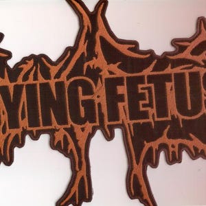 May include: A brown and orange patch featuring the text "DYING FETUS" in bold, dark brown letters. The patch has an irregular, organic shape with a textured, flame-like border in orange.