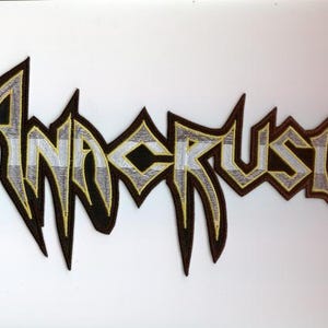 Pode incluir: Patch bordado com o nome da banda "Anacrusis" em uma fonte estilizada e irregular. As letras são preenchidas com um padrão xadrez, contornado em amarelo e marrom, sobre um fundo preto.