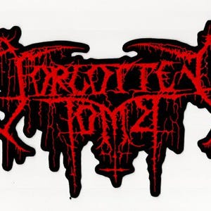 May include: Black and red patch featuring the text "FORGOTTEN TOMB" in a stylized, gothic font. The text is in red with a distressed, cracked effect, set against a black background. The patch has an irregular shape.