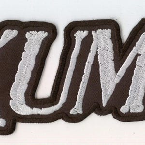 May include: A rectangular embroidered patch featuring the word EXUMER in bold, white, capitalized letters with a textured stitch pattern. The lettering is outlined by a thick, dark brown border that follows the contour of the text.