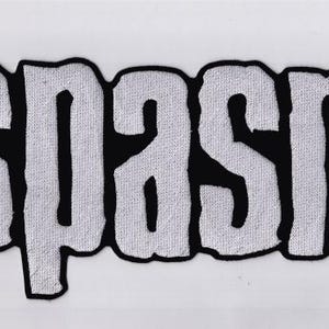 Könnte beinhalten: Ein weißer Aufnäher mit dem Wort "SPASM" in einer kräftigen, strukturierten Schrift. Die Buchstaben sind schwarz umrandet, was einen starken Kontrast erzeugt. Der Aufnäher hat eine leicht verwaschene, Vintage-Ästhetik.