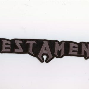 May include: A black and gray patch with the word "TESTAMENT" in stylized lettering. The letters are gray with a black outline, set against a black background. The patch has a jagged edge.