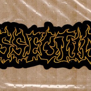 Op de afbeelding: Zwarte patch met gouden, vlamachtige letters. De patch heeft een unieke vorm met afgeronde randen. De tekst lijkt een bandnaam of logo te zijn, met een gotische of metalen esthetiek.