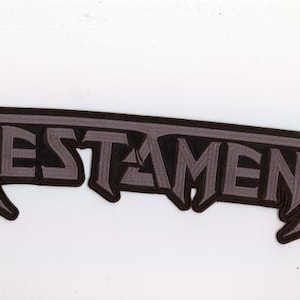May include: A gray and black patch featuring the word "TESTAMENT" in a stylized, gothic font. The patch has a black border and a gray fill, with a slight shadow effect. The design is bold and eye-catching.