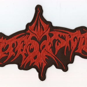 May include: A fabric patch featuring intricate, stylized red embroidery on a dark brown background. The design consists of sharp, jagged, and symmetrical lettering that forms a complex, abstract shape with pointed edges and elongated, flowing lines.