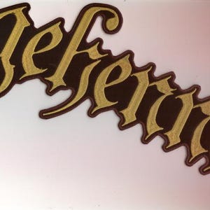 Könnte beinhalten: Ein brauner Aufnäher mit dem Wort "Gefreidig" in goldfarbenen, gotischen Buchstaben. Der Aufnäher hat einen dunkelbraunen Rand und ist vor einem weißen Hintergrund platziert. Die Schriftart ist gotisch.