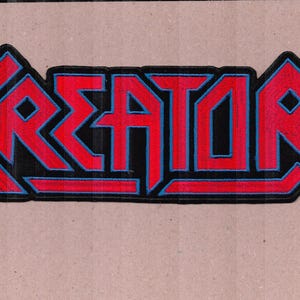 Op de afbeelding: Een geborduurde patch met het woord KREATOR in een gestileerd, hoekig lettertype. De letters zijn felrood met een dunne blauwe rand, geplaatst op een zwarte achtergrond die de contouren van de tekst volgt.