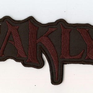 Può includere: Una toppa ricamata rettangolare che presenta la parola KATAKLYSM in un carattere stilizzato rosso scuro con un bordo nero. Le lettere hanno un aspetto strutturato e sono poste su uno sfondo scuro che segue il contorno delle lettere.