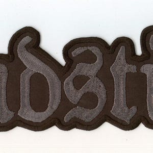 Puede incluir: Un parche bordado de color marrón oscuro que presenta la palabra Endstille en una fuente estilizada de inspiración gótica. Las letras están hechas con hilo gris claro, creando un contraste marcado sobre el fondo de tela marrón oscuro.