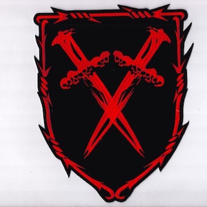 May include: A black shield-shaped patch with a red border and accents. Two crossed red swords with skull-shaped hilts are centered on the patch. The border has a barbed wire design at the top and pointed edges.