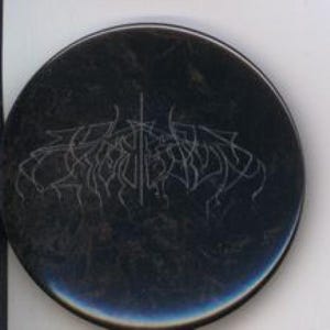 May include: A black, circular button with a metallic sheen. The button features a stylized, white, gothic-style text design. The design is centered on the button and is surrounded by a dark, textured background.