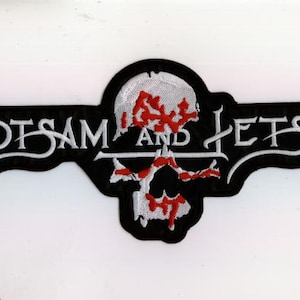 Könnte beinhalten: Schwarz-weißer, gestickter Aufnäher mit den Worten "FLOTSAM AND JETSAM" in stilisierten weißen Buchstaben. Eine Totenkopf-Grafik mit roten, blutartigen Akzenten ist mittig auf dem Aufnäher platziert.