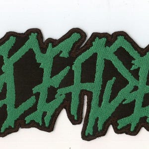 May include: A rectangular embroidered patch featuring the word DECEASED in a jagged, stylized green font with a black border. The lettering has a distressed, sharp appearance, and three small green dots follow the text at the bottom right.