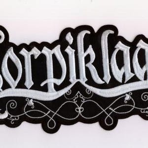 以下が含まれることがあります： 黒い背景に白いゴシック体で「Korpiklaani」と刺繍されたパッチ。パッチには装飾的な白い縁取りと、下部に様式化されたデザインがあります。
