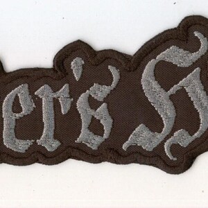 Könnte beinhalten: Ein rechteckiger gestickter Aufnäher mit dem Text Master's Hammer in einer stilisierten gotischen Schriftart. Die Schrift ist silbergrau mit einem dunkelbraunen Rand, der der Kontur der Wörter folgt und ein markantes visuelles Erscheinungsbild erzeugt.