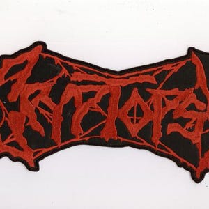 May include: A black patch with red embroidered text. The text is stylized and appears to be a band logo. The patch has a unique shape with a jagged edge.