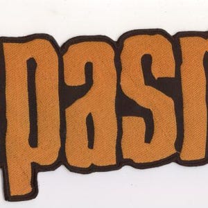 May include: An orange and yellow patch with the word "SPASM" in large, stylized letters. The letters are outlined in dark brown, creating a bold contrast against the white background. The patch has a textured appearance.