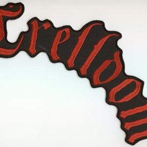 Puede incluir: Un parche negro con la palabra "Freedom" bordada en letras góticas rojas. El parche tiene una forma ondulada e irregular y un borde negro. El texto está en una fuente estilizada y antigua.