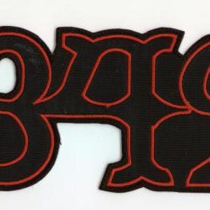 Op de afbeelding: Zwarte patch met het nummer 1349 in een opvallend, gestileerd lettertype. De cijfers zijn zwart met een rode omlijning, wat een opvallend contrast creëert. De patch heeft een getextureerd uiterlijk, mogelijk gemaakt van stof of een vergelijkbaar materiaal.