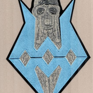 May include: Embroidered patch featuring a stylized skull design in gray and black, set against a light blue geometric shape with a black border. The patch has a detailed, textured appearance, suitable for jackets or bags.