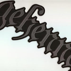 Könnte beinhalten: Schwarzer Aufnäher mit dem Wort "Gefang" in gotischer Schrift. Die Buchstaben sind grau mit schwarzem Umriss und schwarzem Hintergrund. Der Aufnäher befindet sich auf weißem Hintergrund.