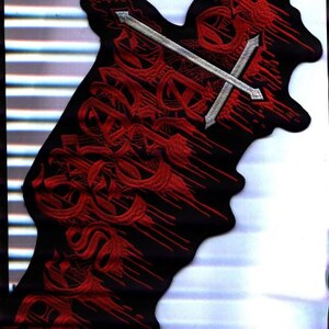 Peut inclure: Écusson noir brodé avec un motif rouge comprenant des lettres stylisées, une tête de mort et une croix argentée. L'écusson a une forme unique et est probablement destiné aux vêtements ou accessoires.