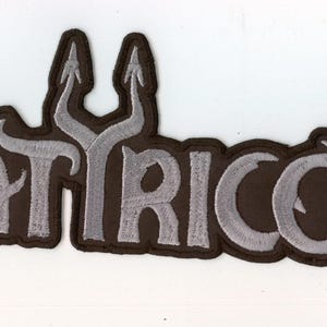 以下が含まれることがあります： 鋭いエッジの効いたスタイリッシュなフォントで「SATYRICON」という文字が刺繍されたワッペン。文字はライトグレーで、ダークブラウンの縁取りが施されており、コントラストの強いデザインと角張った文字の美しさが特徴的です。