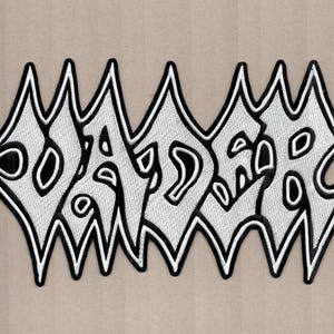 以下が含まれることがあります： 「DADDER」の文字が、ギザギザとした書体で刺繍されたパッチ。テキストは白で黒い縁取りがあり、ニュートラルな背景に配置されています。このパッチは、ユニークでエッジの効いたデザインです。