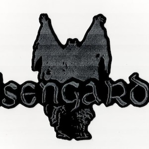 Pode incluir: Patch preto Isengard com um design estilizado. O patch apresenta a palavra "Isengard" em uma fonte gótica, com uma silhueta escura e texturizada acima e abaixo do texto. O design está em um fundo branco.
