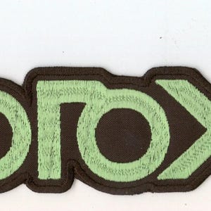 Könnte beinhalten: Ein gestickter Aufnäher mit stilisierten, kantigen Buchstaben in hellminzgrünem Garn mit dunkelbraunem Rand. Der Text lautet "CYGOGOXII" in einer fetten, geometrischen Schriftart, wobei die Buchstaben zu einer Einheit verbunden sind.
