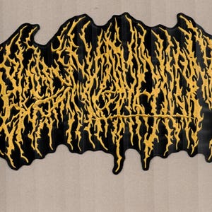 Peut inclure: Écusson brodé noir avec un motif doré en forme de flamme. Le patch a une forme irrégulière et un aspect texturé. La broderie dorée ressort sur le fond noir, créant un contraste saisissant. Idéal pour les vestes, sacs ou autres vêtements.