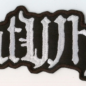 Può includere: Una toppa ricamata nera che presenta la parola Goatwhore in un carattere bianco stilizzato e frastagliato. La toppa ha un bordo nero spesso che segue il contorno delle lettere, creando una forma distinta per questo accessorio in tessuto.