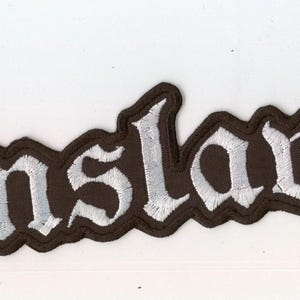 May include: An embroidered patch featuring the word Enslaved in a stylized white gothic font with a thick black border. The lettering has a textured appearance created by dense white thread stitching against a solid black fabric background.