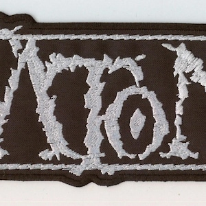 Könnte beinhalten: Ein rechteckiger, bestickter Aufnäher mit schwarzem Hintergrund und dem Wort KATATONIA in einer zackigen, silberfarbenen Schrift. Der Text ist von einem dünnen, silbernen rechteckigen Rahmen umgeben, und der Aufnäher hat einen schwarzen Rand.
