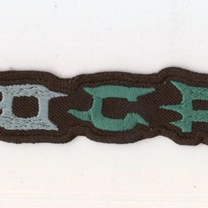 Peut inclure: Un écusson brodé rectangulaire affichant le mot HYPOCRISY dans une police stylisée et dentelée. Les lettres sont un mélange de gris clair et de vert sarcelle, sur un fond marron foncé avec une bordure épaisse de la même couleur.