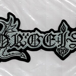 Könnte beinhalten: Gestickter Aufnäher mit dem Wort "Hordes" in gotischer Schrift, schwarz umrandet und hellgrau gefüllt. Der Aufnäher hat einen schwarzen Rand und dekorative Elemente an jedem Ende.