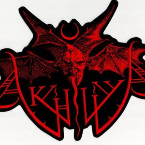 Op de afbeelding: Zwart en rood embleem met een demonisch gezicht, vleermuisvleugels en het woord "AKHV". Het ontwerp bevat een halve maan en een lange, gespleten tong. Het embleem heeft een unieke, edgy esthetiek.