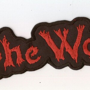 Op de afbeelding: Een zwarte geborduurde patch met de rode tekst "In the Woods..." in een gestileerd, grillig lettertype. De patch heeft een donkere rand die de contouren van de letters volgt, wat een opvallende vorm geeft aan dit stoffen accessoire.