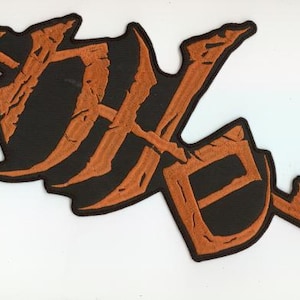 May include: A black patch featuring an orange, stylized design. The design is a combination of letters and shapes, with a textured, distressed look. The patch is set against a white background.