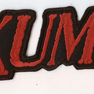 Könnte beinhalten: Ein rechteckiger gestickter Aufnäher mit dem Wort EXUMER in fetten, roten Großbuchstaben mit einem dicken schwarzen Rand. Die Schrift hat ein strukturiertes, gesticktes Aussehen und das gesamte Design ist auf einem einfarbigen schwarzen Hintergrund.