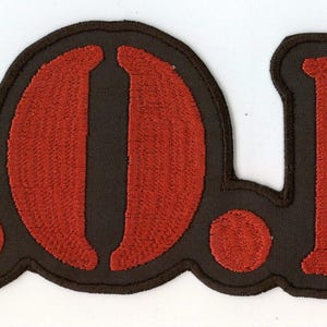 Può includere: Una toppa ricamata che presenta le lettere S.O.D. in filo rosso brillante su sfondo nero. Le lettere sono stilizzate con un carattere spesso e arrotondato, e l'intera toppa è bordata di nero, creando un design distintivo ad alto contrasto.