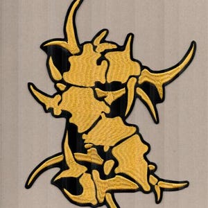 Op de afbeelding: Geborduurde patch met een abstract goud en zwart ontwerp. De patch heeft een getextureerde uitstraling met een complex, gelaagd patroon. Het ontwerp bevat meerdere gebogen, hoornachtige vormen. De patch is waarschijnlijk voor kleding of accessoires.
