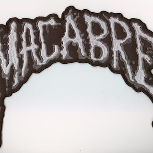 Puede incluir: Un parche bordado que presenta la palabra MACABRE en una fuente estilizada y goteante. Las letras son blancas con un contorno gris oscuro, sobre un fondo marrón oscuro que sigue la forma irregular y orgánica del texto.