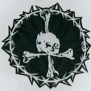 Puede incluir: Parche negro con un diseño de calavera y huesos cruzados blancos, rodeado por una corona de alambre de púas. El parche tiene forma circular con bordes dentados, creando una estética gótica audaz.