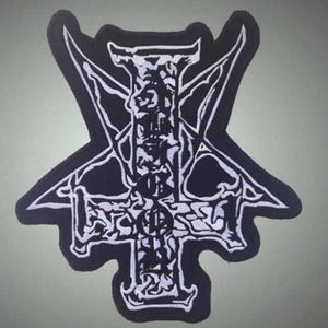 May include: Black and white embroidered patch featuring a stylized cross design with intricate details. The patch has a unique shape with pointed edges and a border. Ideal for jackets, bags, or other apparel.
