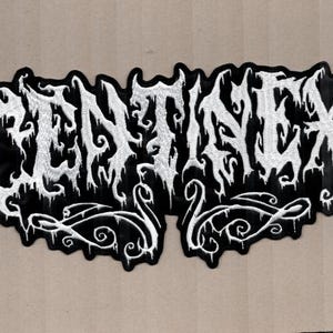 May include: A black and white embroidered patch with the word "DENTINEX" in a gothic font. The patch has a black background with white lettering and decorative flourishes.