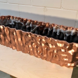May include: A long, copper-colored planter with a textured, wavy surface. The planter has a black interior and a divider in the middle.