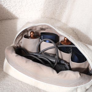 May include: A cream-colored zippered cosmetic bag with a quilted interior, holding hair styling tools. The bag contains a hair dryer, brush, and other accessories. The bag is open, revealing the contents.